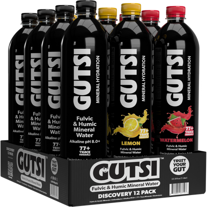 A 12-pack display of GUTSI Fulvic & Humic Mineral Water bottles in a black cardboard tray. The front bottle is Lemon, with Watermelon and other flavors visible behind it.