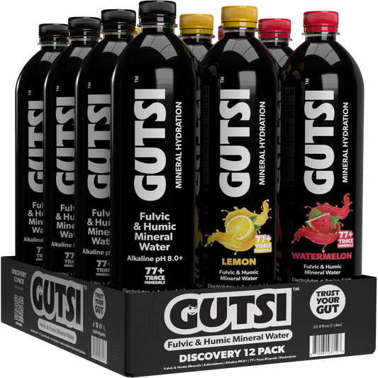 A 12-pack display of GUTSI Fulvic & Humic Mineral Water bottles in a black cardboard tray. The front bottle is Lemon, with Watermelon and other flavors visible behind it.