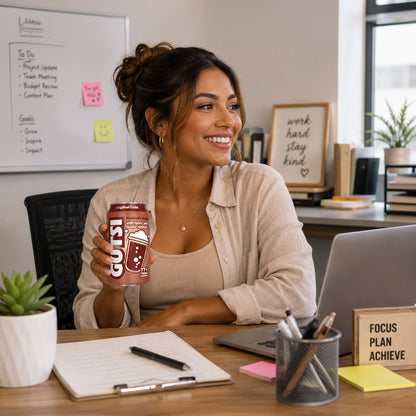 A woman in a beige shirt sits at a desk in a bright office, holding a can of Gutsi Original Cola Soda. The desk is cluttered with a laptop, notebook, pens, and sticky notes, and a whiteboard is visible in the background.