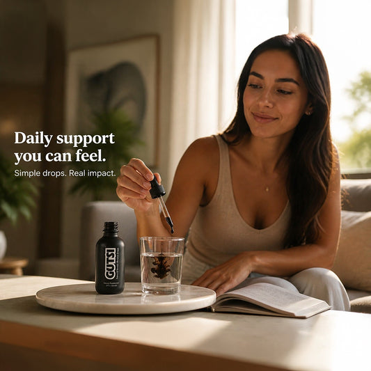 A woman in a beige tank top sits on a couch in a living room, holding a dropper above a glass of water. A black bottle labeled GUTSI sits on a small round table beside her.