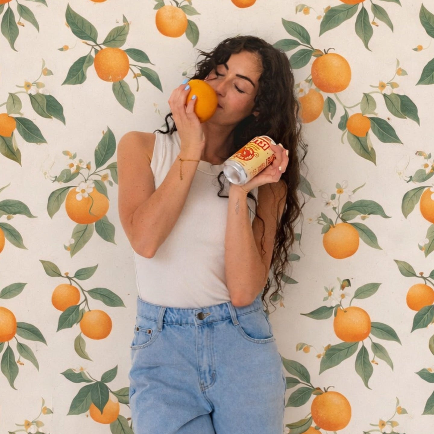 A woman with long dark curly hair poses against a cream wallpaper patterned with oranges and green leaves, holding an orange to her lips and a can of GUTSI Orange Cream soda in one hand.